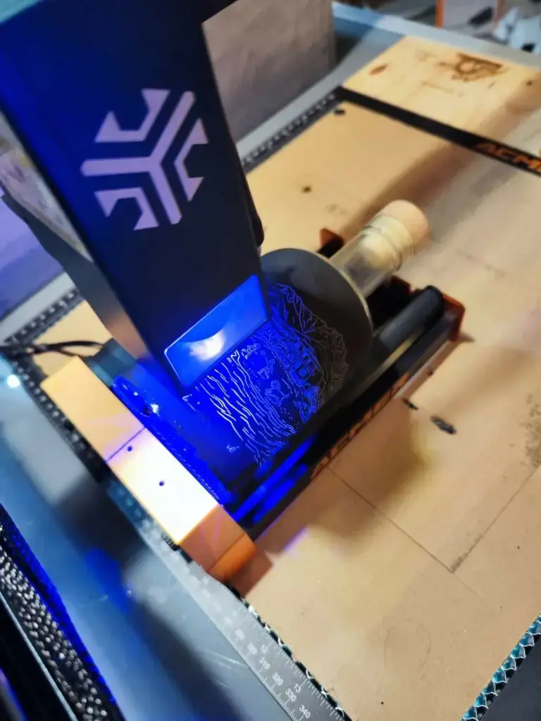 Gravure laser diode en cours sur bouteille en verre avec accessoire rotatif — laser ACMER, faisceau bleu 455 nm visible, primaire noir Le Graveur™️appliqué sur la surface