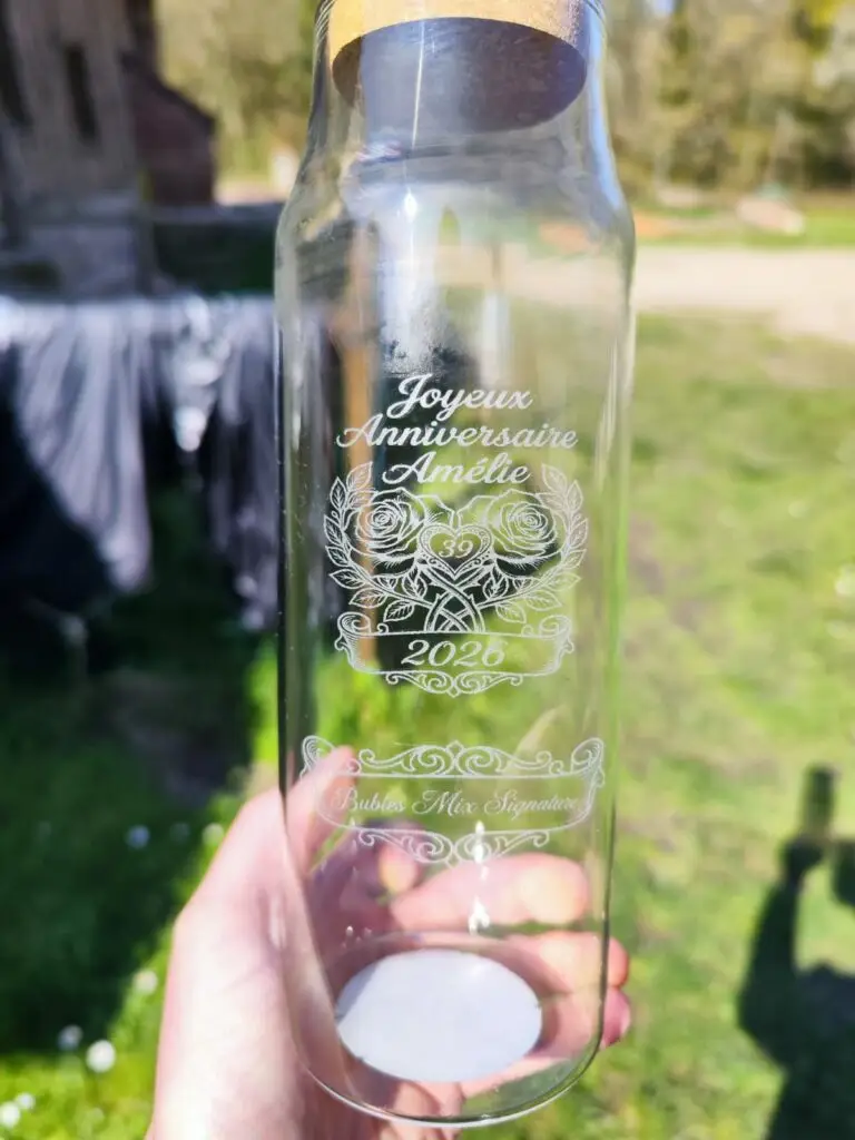 Bouteille en verre gravée au laser diode tenue en plein soleil — motif floral détaillé et texte "Joyeux Anniversaire Amélie 39 2026" en script calligraphique, gravure blanche sur verre transparent