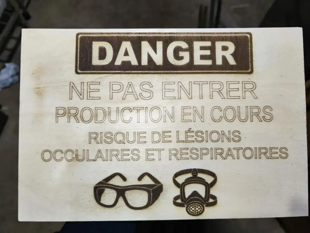 Panneau de sécurité gravé au laser sur bois — "DANGER Ne pas entrer, production en cours, risque de lésions oculaires et respiratoires" avec pictogrammes lunettes et masque respiratoire