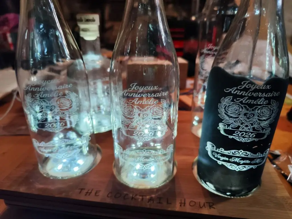 Trois bouteilles en verre gravées au laser diode — motif floral et texte "Joyeux Anniversaire Amélie 2026", présentées sur plateau bois gravé "The Cocktail Hour"