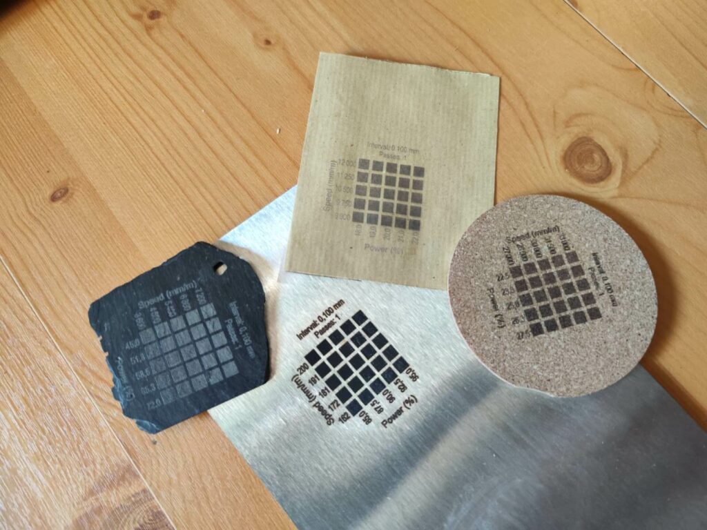Quatre grilles de test laser réelles gravées sur papier kraft, ardoise noire, liège et inox, matrices vitesse × puissance obtenues avec un graveur laser diode